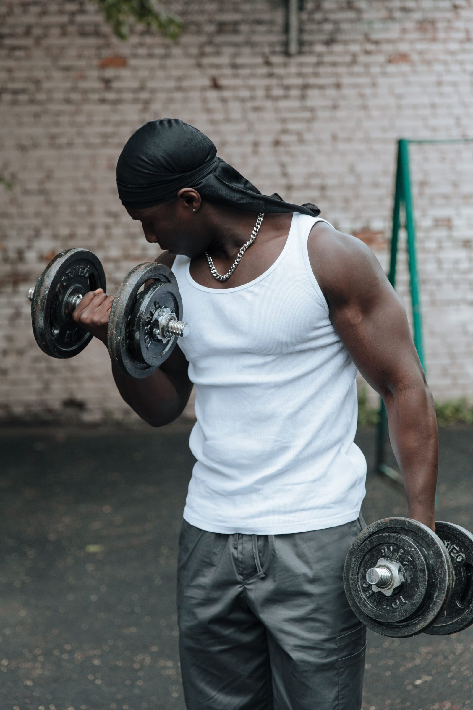 An adult man lifts dumbbells outdoors wearing a tank top and headscarf.
