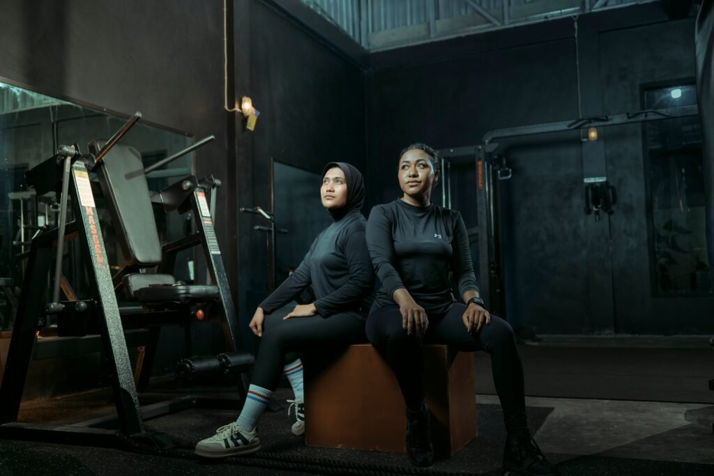 Two women in athletic wear sitting on a box with gym equipment in the background.